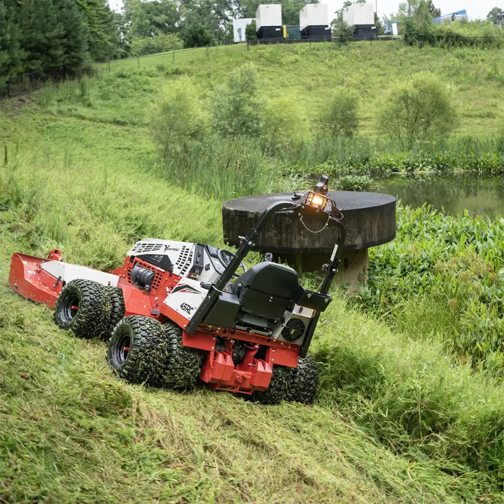 A remote-control slope mower cutting tall grass on a steep bank beside a pond